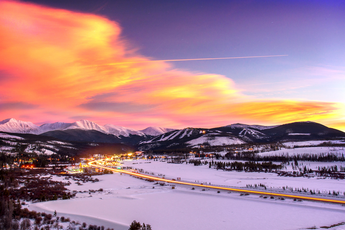 A panoramic image of the mountains of Winter Park, Colorado, under a setting-sun sky, paintedin neon, wispy orange clouds. There is snow everyone on the ground and the lights from the city glimmer in the night sky.