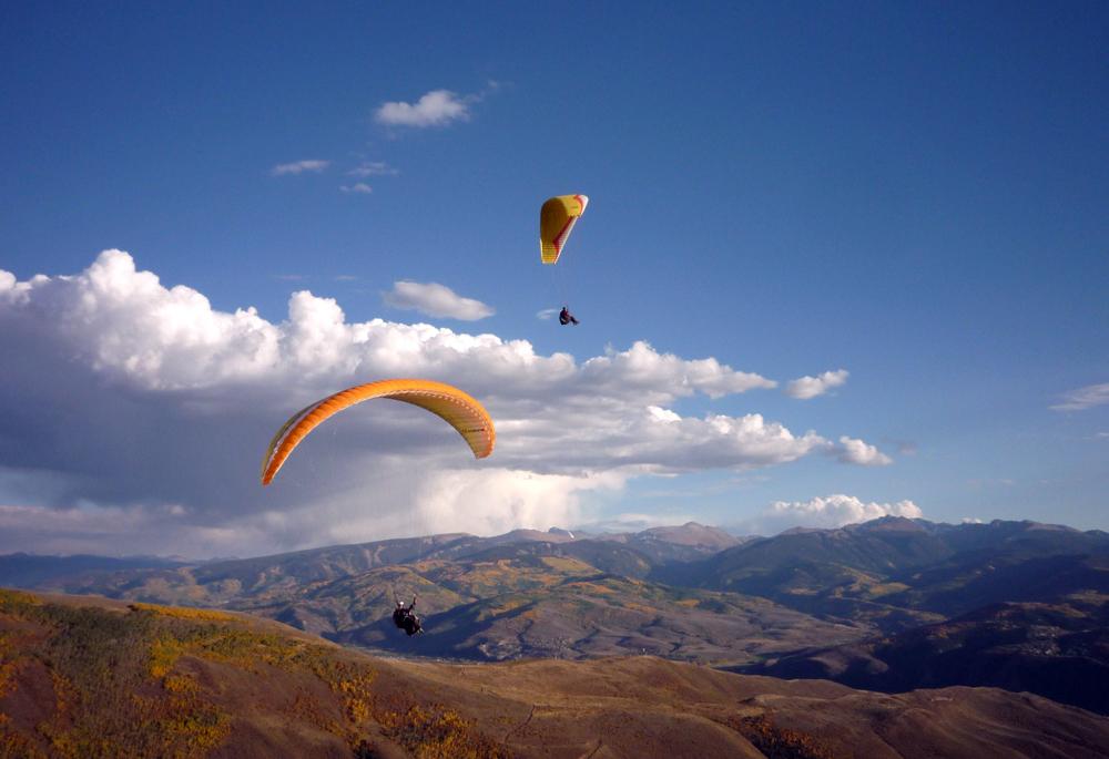 Two hang gliders glide through the blue sky and white clouds over a mountain range in the fall.